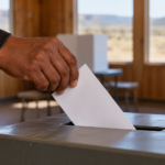 A hand places a ballot in a ballot box, with voting booths and a desert landscape in the background.